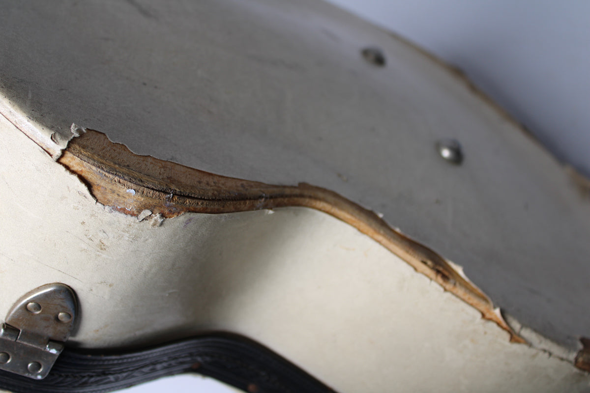 Close-up of the tan archtop case's worn edge, revealing exposed wood and peeling material, with a silver hinge visible at bottom left.
