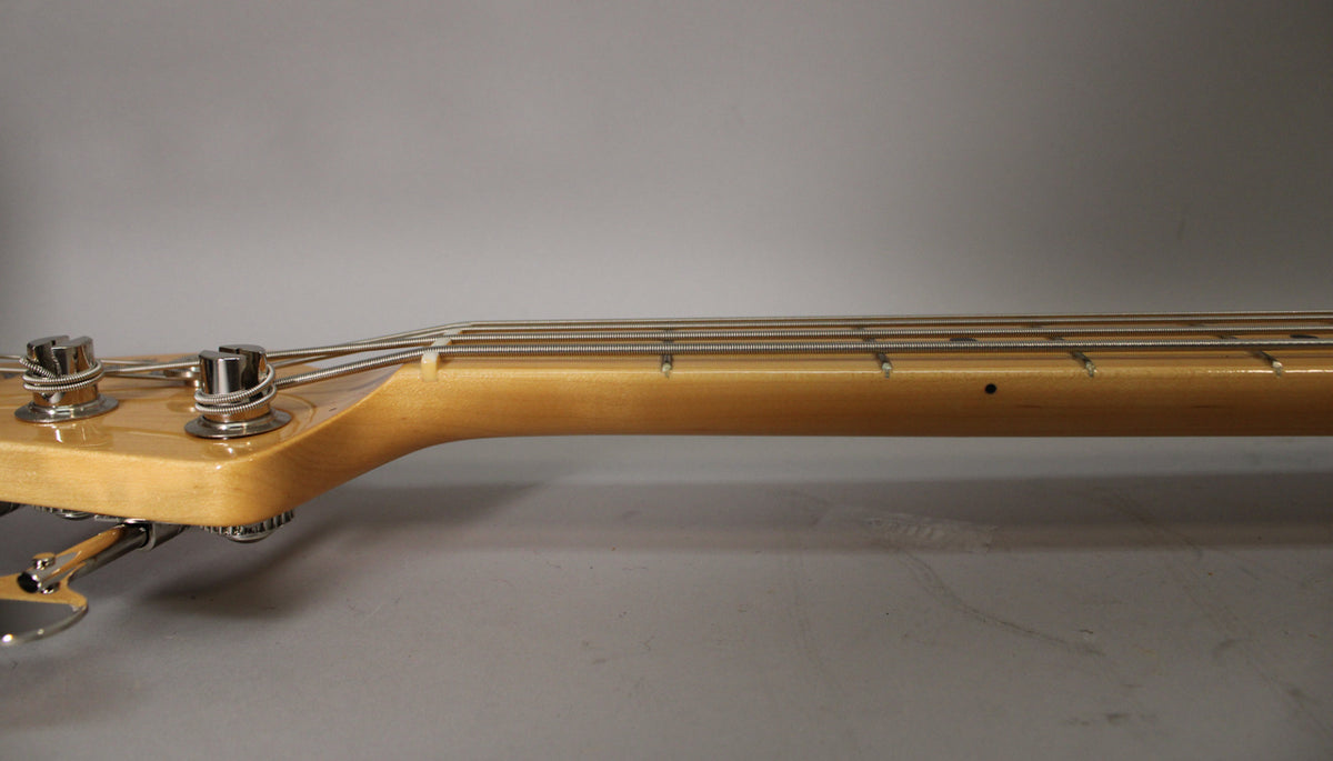 Close-up of the bass neck and headstock from the side, showing the light maple finish, chrome tuners, and string tree.