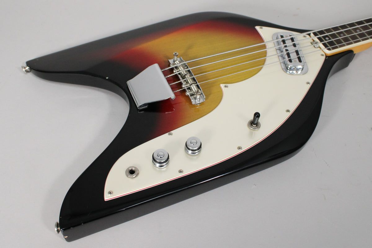 Angled view of the sunburst bass body, showing the white pickguard with red trim, chrome bridge cover, two knobs, and a toggle switch.