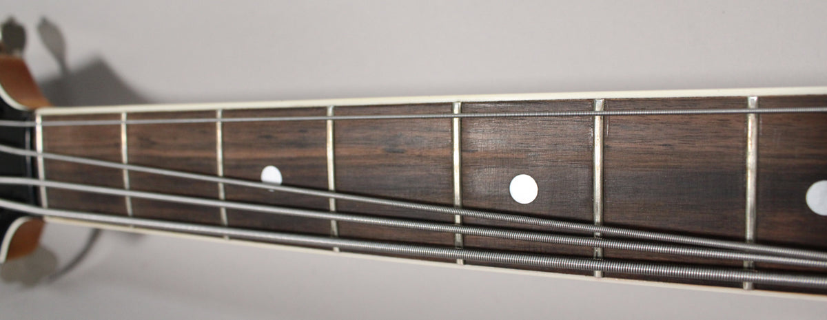 Close-up of the bass fretboard, showing dark wood, metal frets, white dot inlays, and four strings with visible winding.