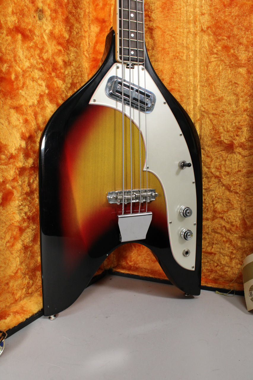 The bass's sunburst body with a white pickguard, chrome hardware, and three control knobs is angled in its orange-lined case.
