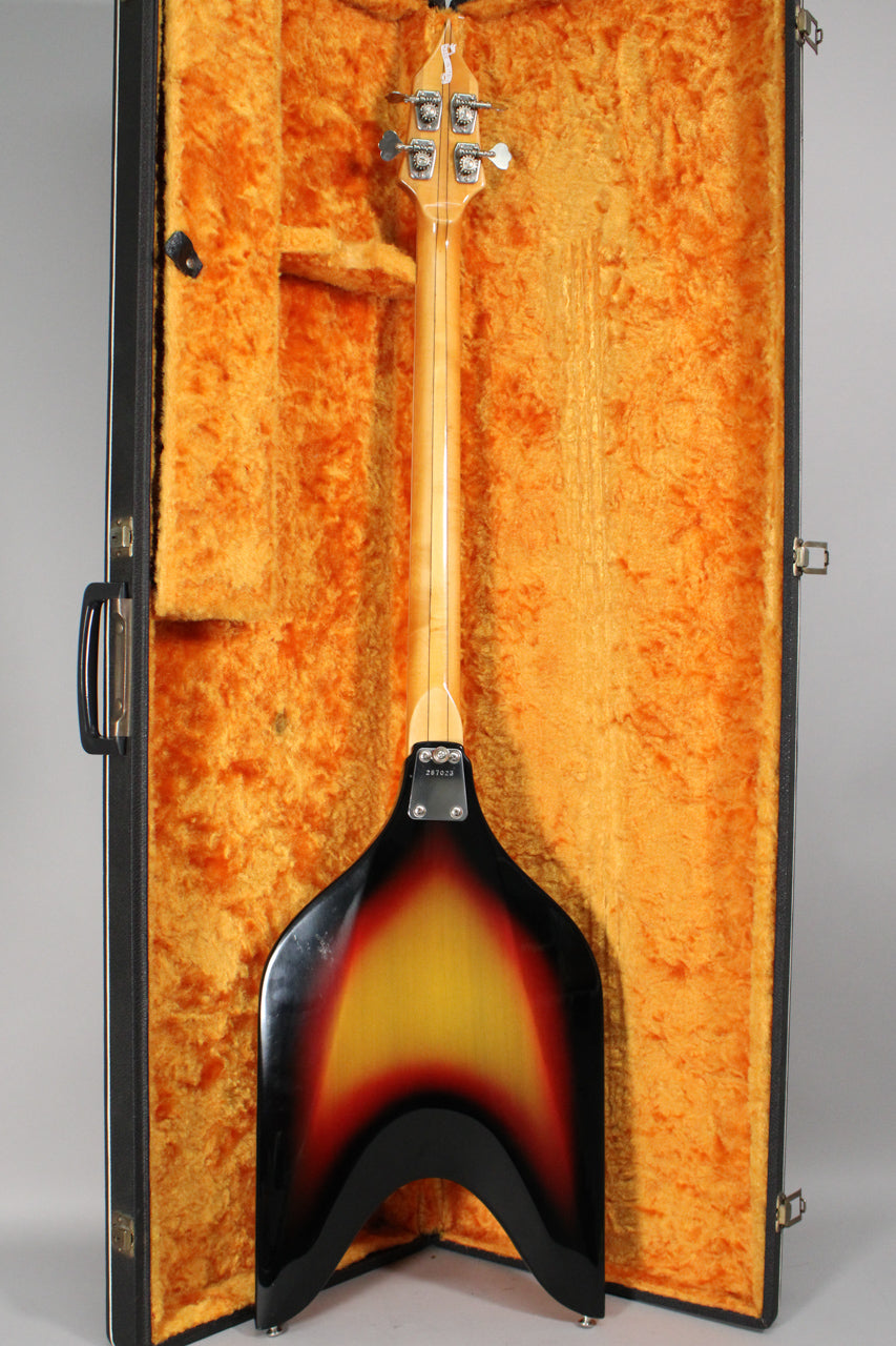 The bass is shown from behind in its open, orange-lined case, revealing the sunburst V-shaped body and light wood neck.