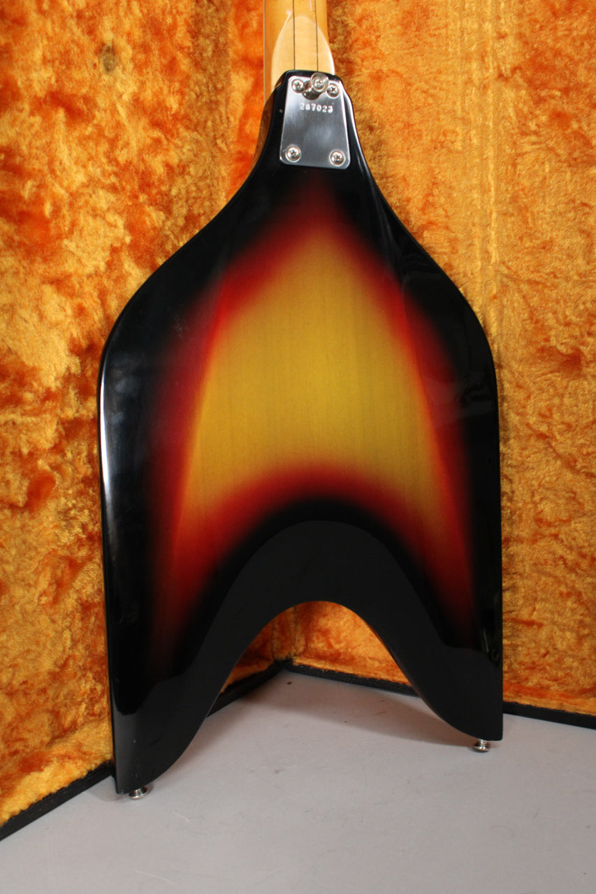 The bass's V-shaped sunburst body, with a black-red-yellow gradient, is seen from the back, showing the neck plate and strap buttons.