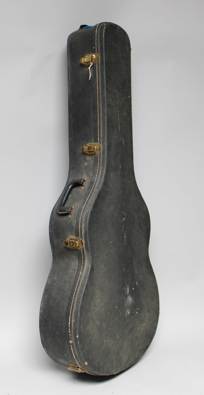 A black hollowbody guitar case stands upright, showing its worn, textured surface, three brass latches, and a black handle.