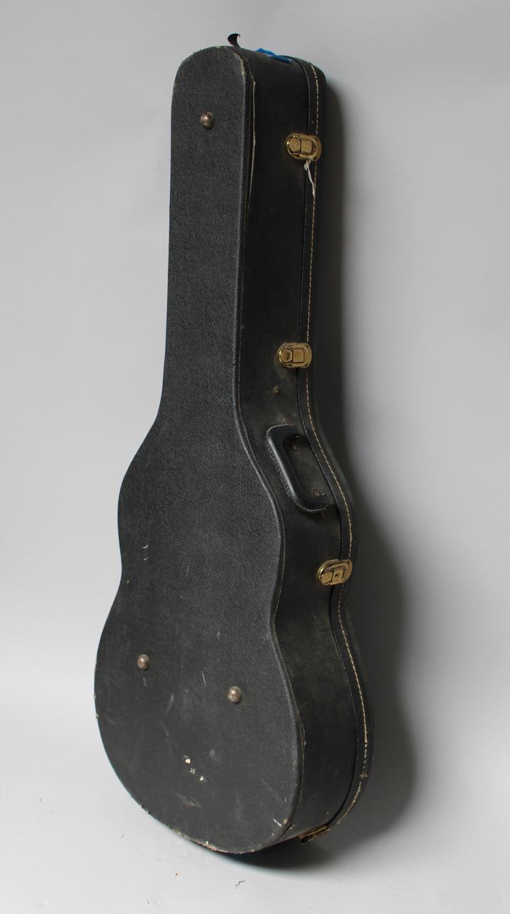 A black textured guitar case stands upright, angled to show its side with three brass latches, a handle, and worn edges.