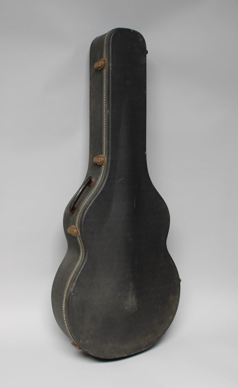 A black guitar case stands upright, showing its side with white stitching, three tarnished latches, and a worn handle.