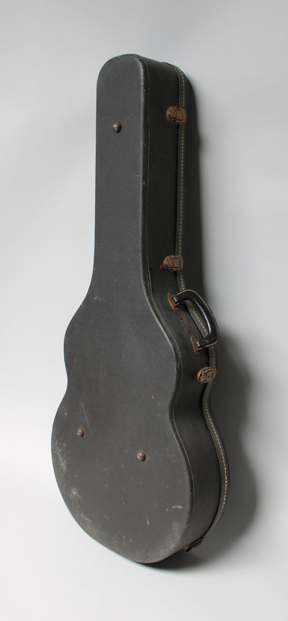 A black guitar case stands upright, showing its side profile with a handle and two latches, revealing wear and scuffs.