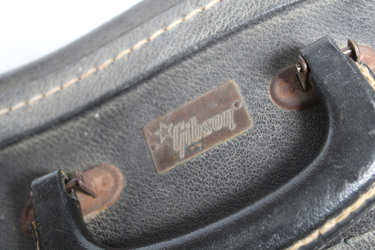Close-up of the black textured case, showing the worn handle, rusty hardware, and a metal badge with a star and "Gibson" text.