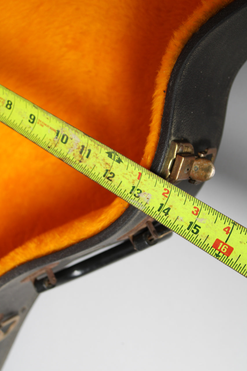 A bright yellow tape measure extends across the open black case, showing the orange plush lining and a tarnished brass latch.