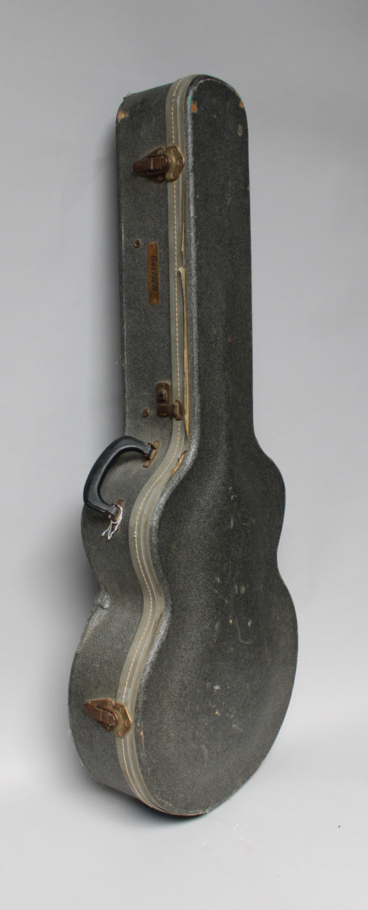 A vintage, dark gray, textured guitar case stands upright, showing its side profile with a black handle and brass latches.