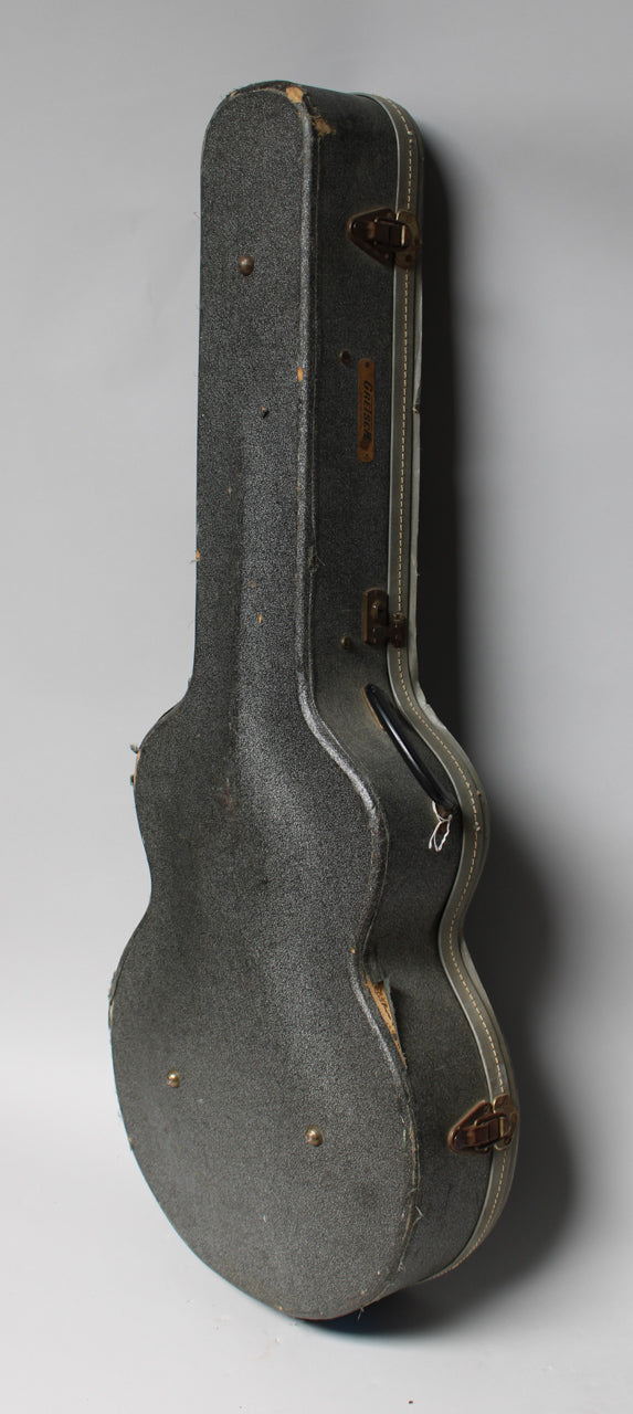 The guitar case stands upright, showing its dark gray textured exterior, worn edges, and a brown handle with a metal latch.