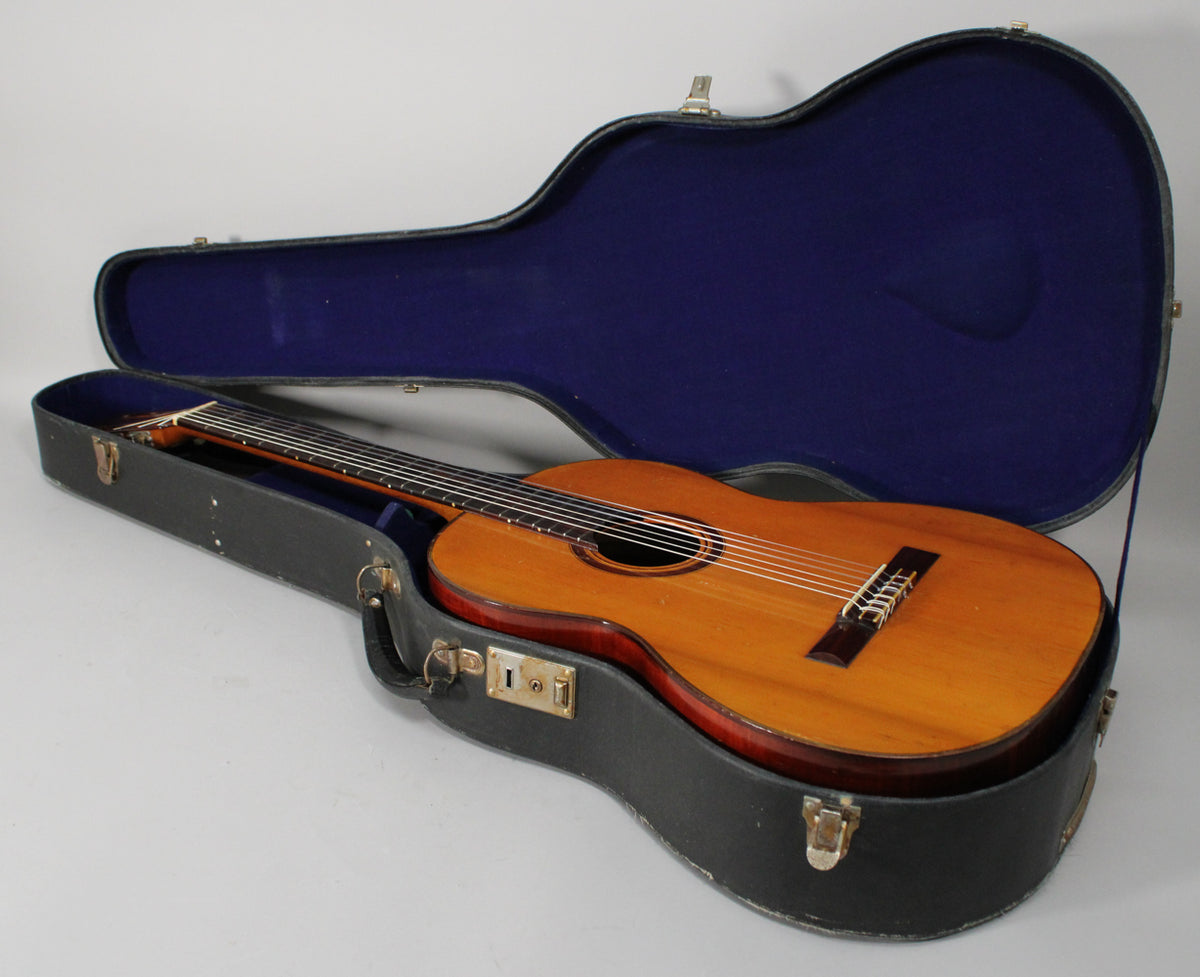 The classical guitar with a light brown top and dark reddish-brown sides rests in its open black hard case with a dark blue plush interior.