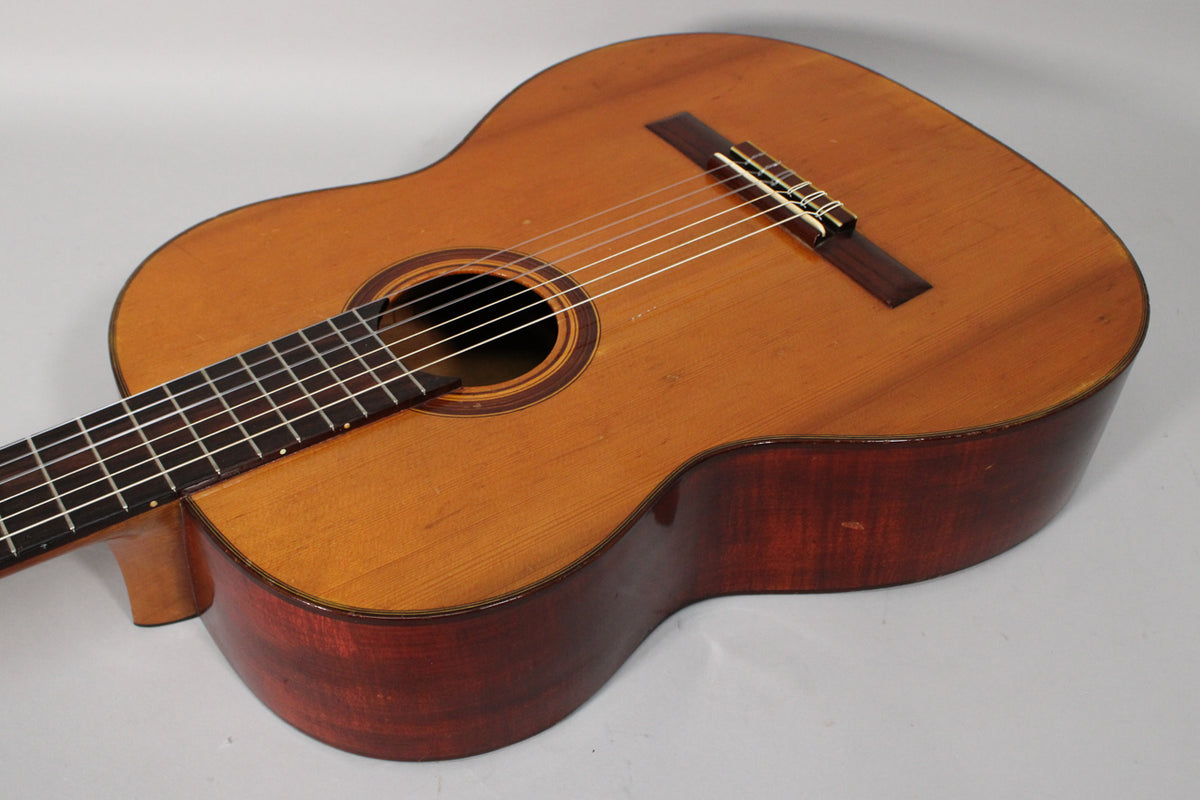 The classical guitar's light brown top and dark reddish-brown sides show light wear, with a dark fretboard and bridge.