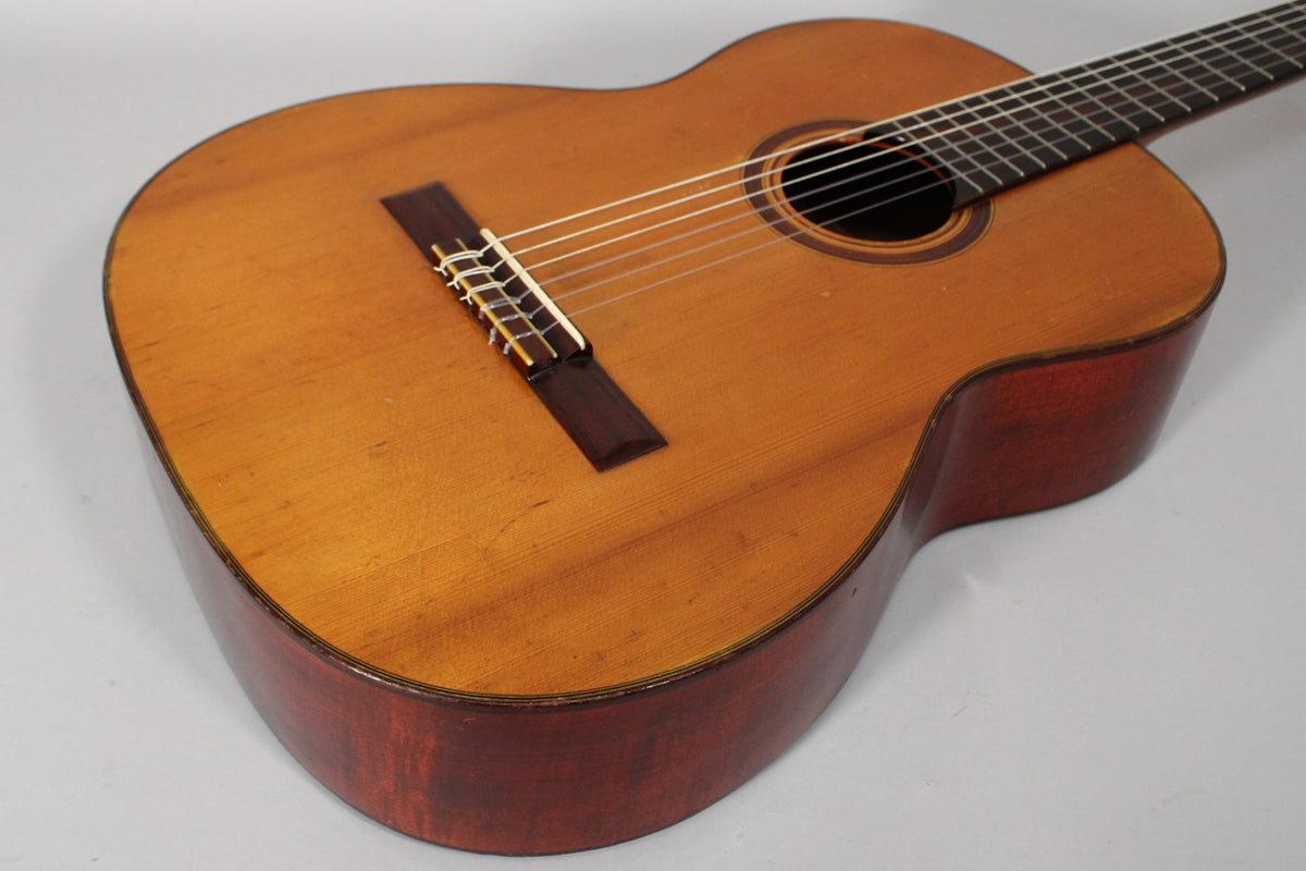 Overhead view of the classical guitar, showing the light brown top with a dark soundhole rosette and reddish-brown sides, with wear.