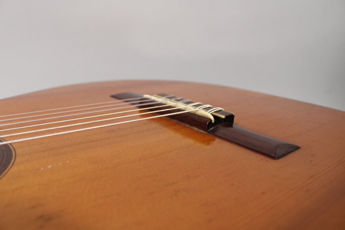 Close-up of the classical guitar's light brown body, showing the dark brown bridge, white saddle, and six white strings.