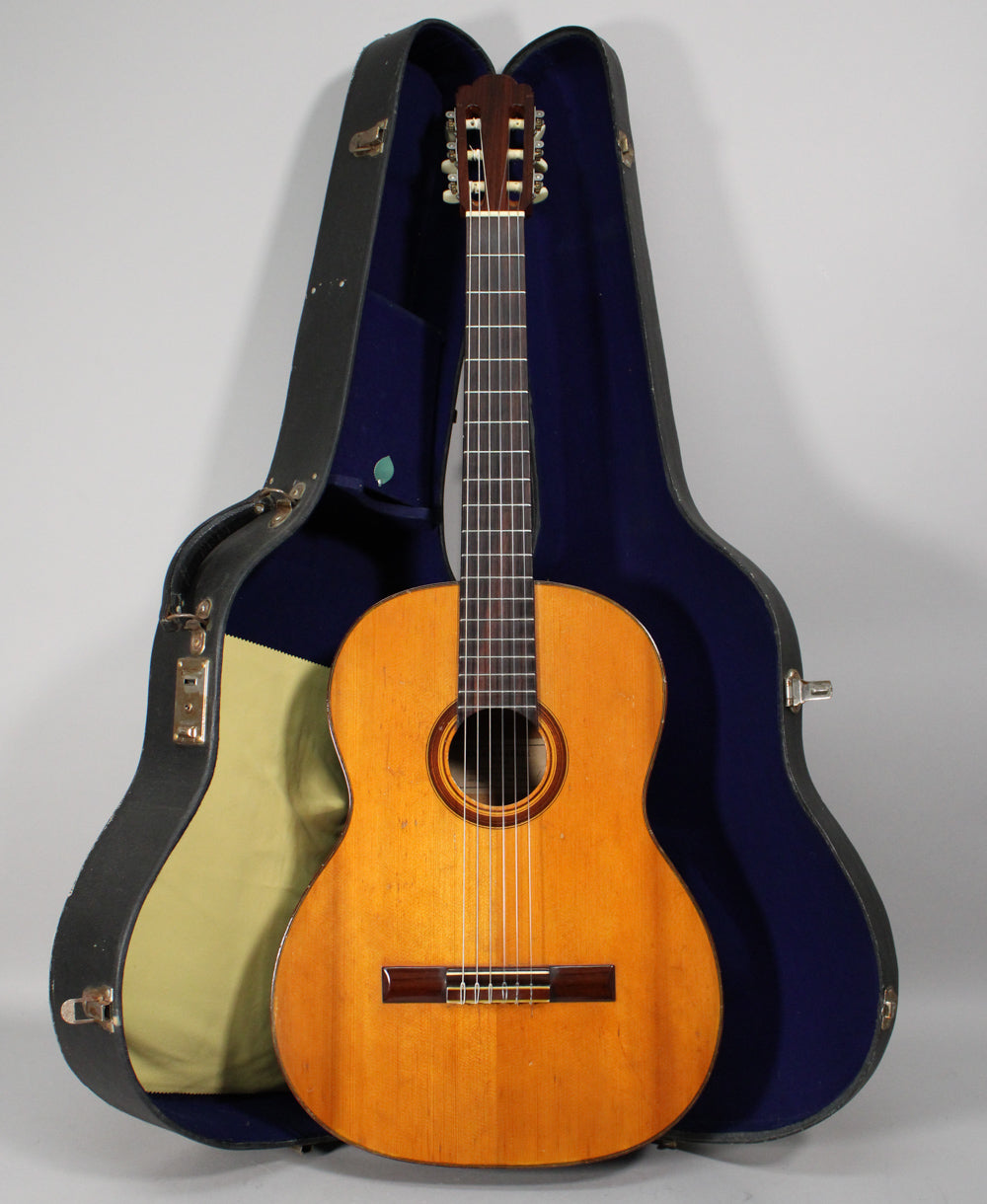 Full-front view of a light-brown classical guitar with dark fretboard and bridge, standing in an open, dark-blue lined hard case.