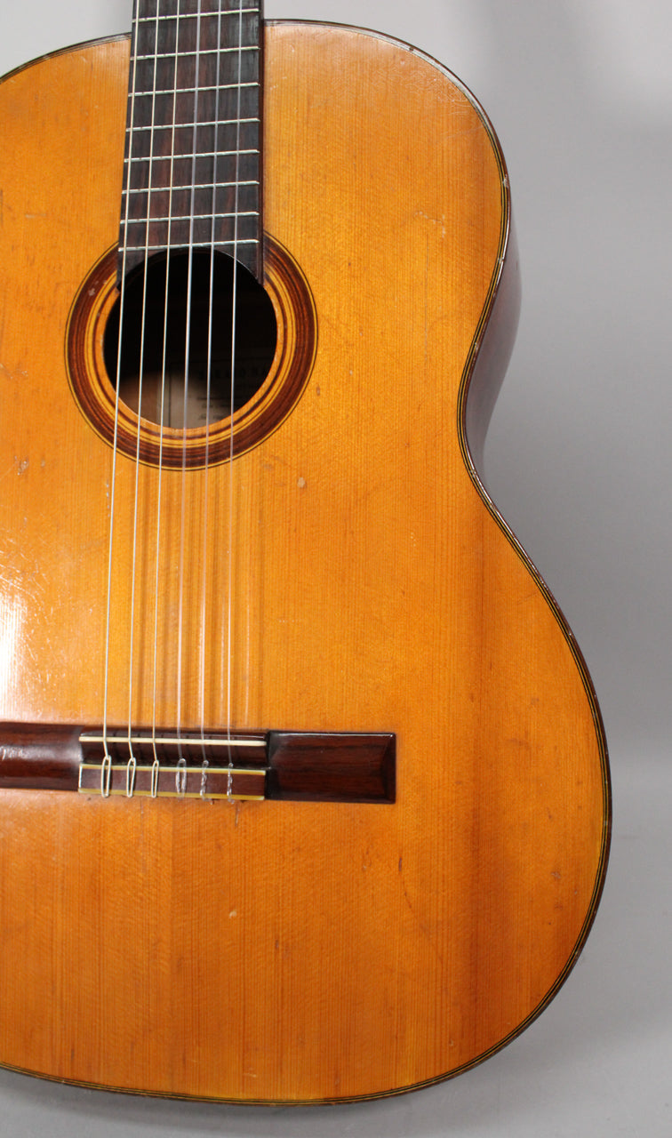 The classical guitar's light-colored body shows visible wear, scratches, and finish checking, with a dark wood bridge and rosette.