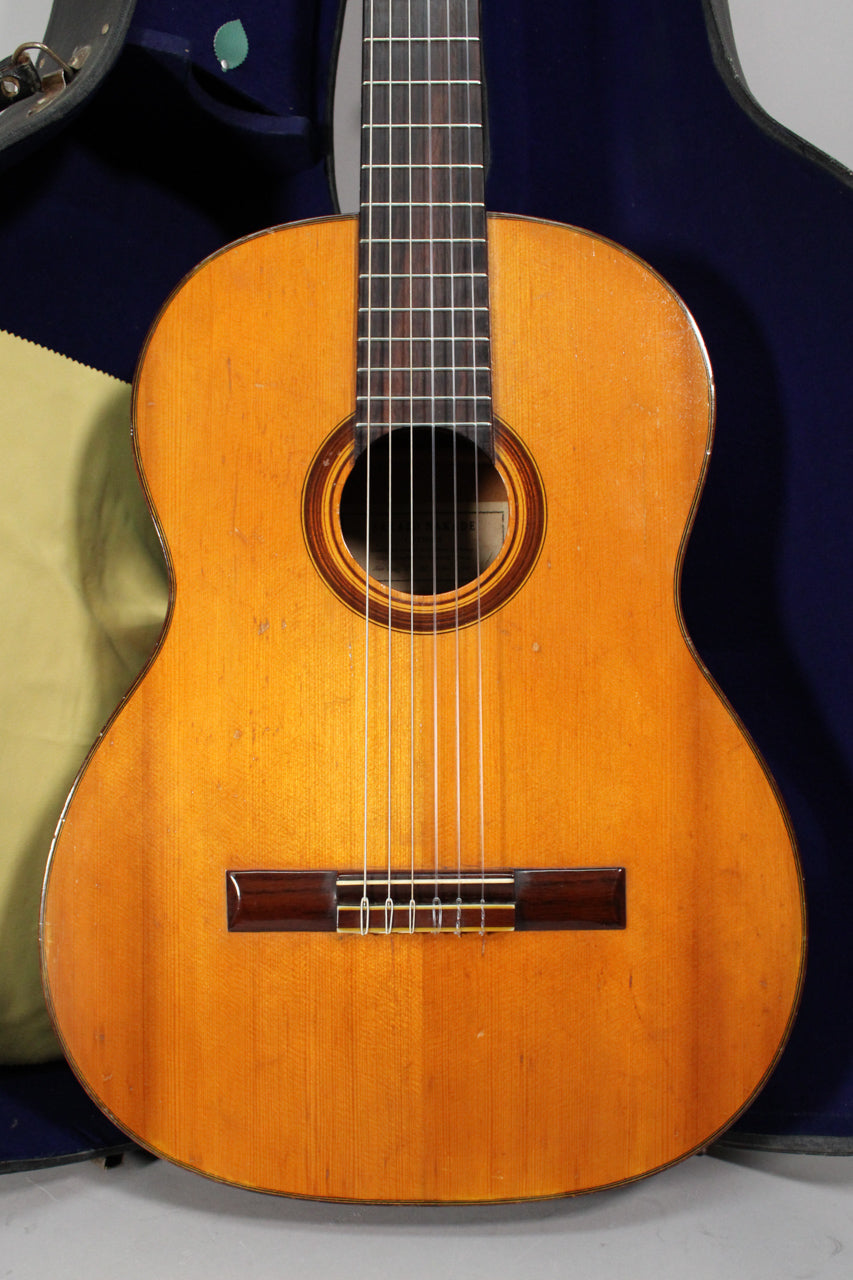 The classical guitar's light-amber body and dark fretboard are framed by a blue case, showing some finish wear and checking.