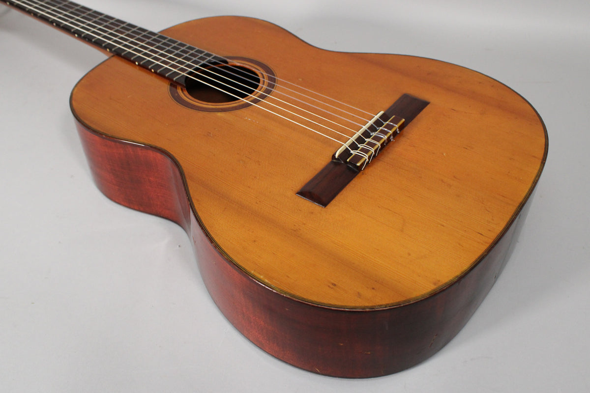 The classical guitar's light-brown top and dark-red sides show visible wear, with a dark wood bridge and white saddle.