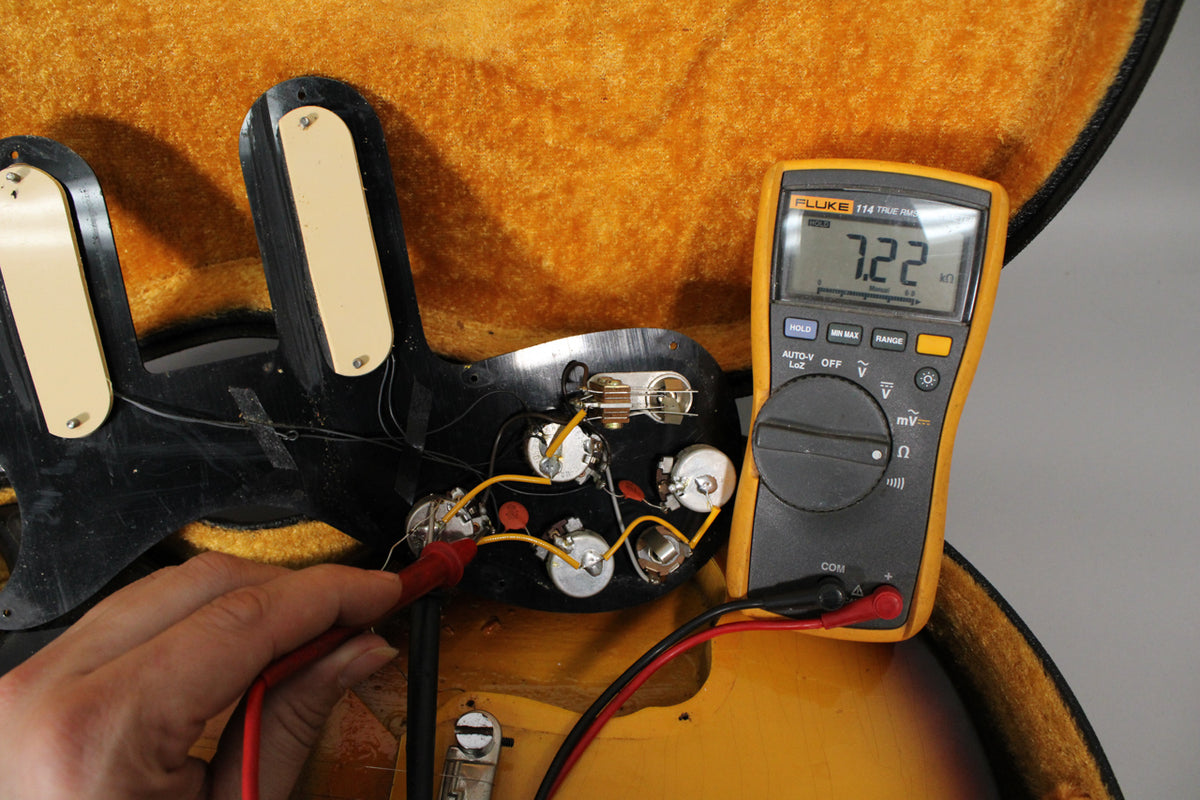 A hand holds a multimeter probe to the guitar's exposed electronics, showing a 7.22 kΩ reading on the display.