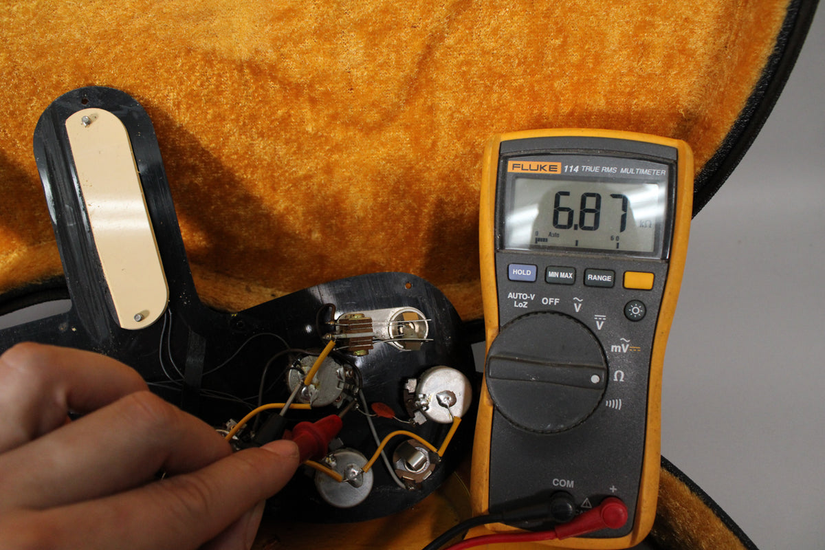 A hand tests the guitar's exposed electronics with a multimeter reading 6.87, showing the wiring and pots against a plush case lining.