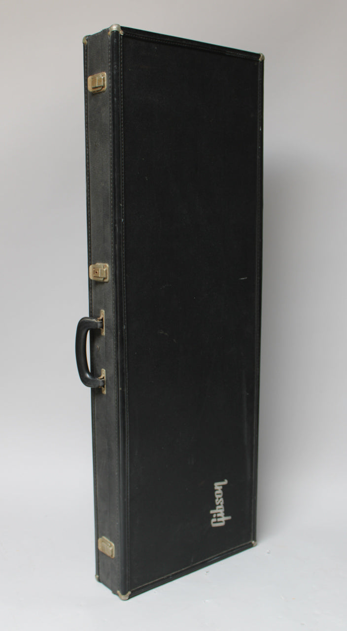 A black guitar case stands upright, showing its side with a handle, two gold latches, and worn metal corner protectors.