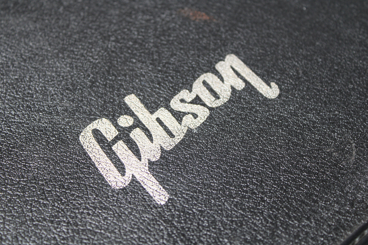 Close-up of the black textured case with the white Gibson logo, showing cracking and wear on the logo and a reddish mark above it.