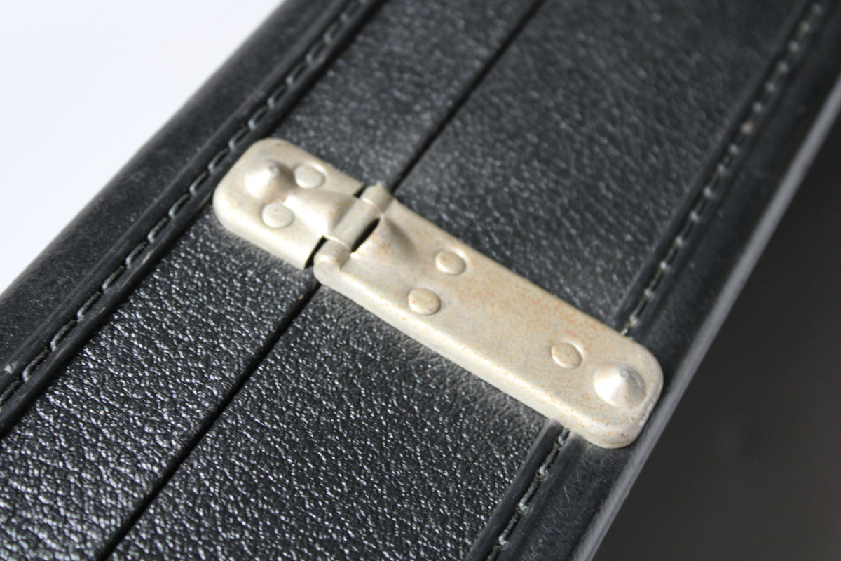 Close-up of a tarnished, silver-colored metal hinge on the black textured exterior of the case, showing wear and light rust.