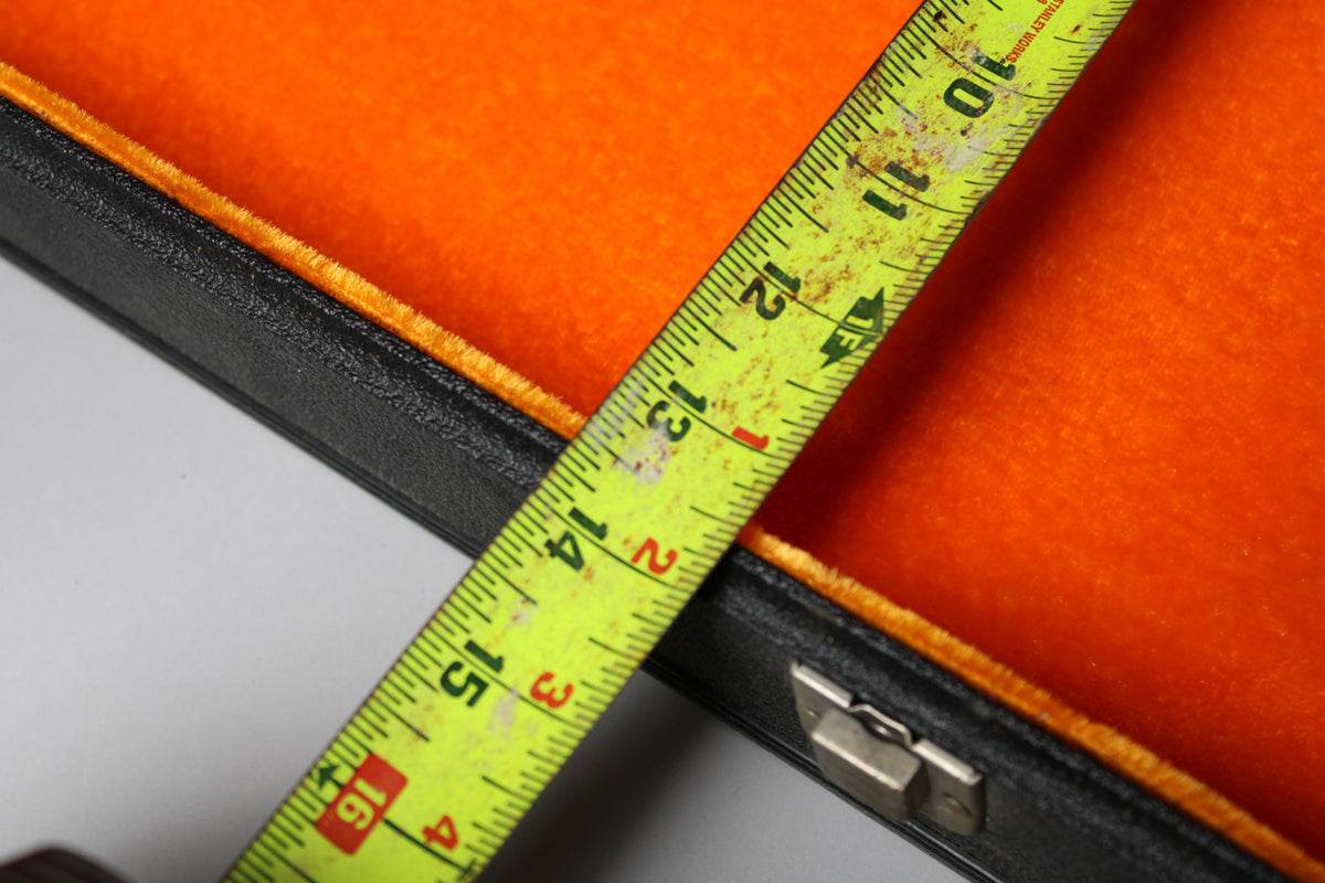 A close-up of the open black case with orange plush lining, showing a metal latch and a yellow measuring tape across it.