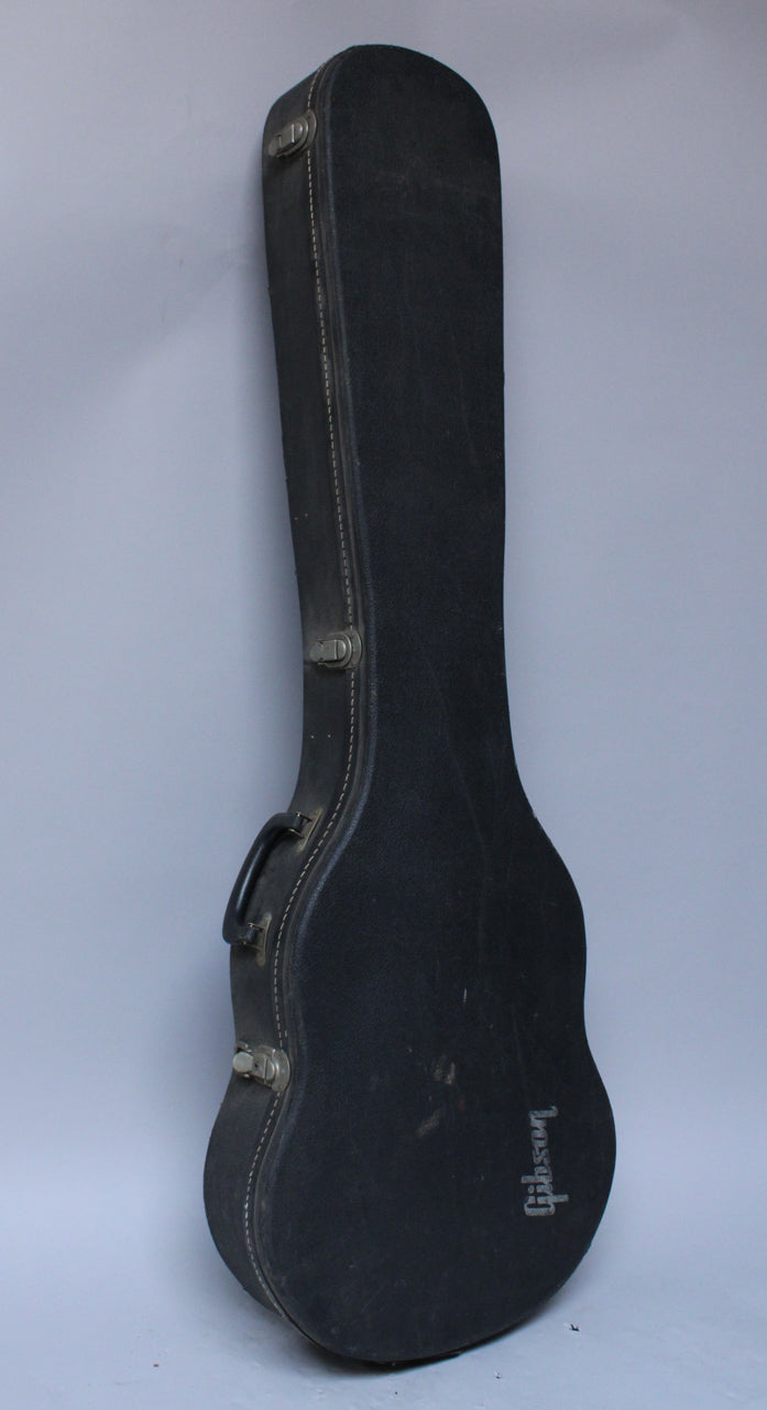 Black guitar case standing upright, showing its side with a handle, three latches, and white stitching along the edge.