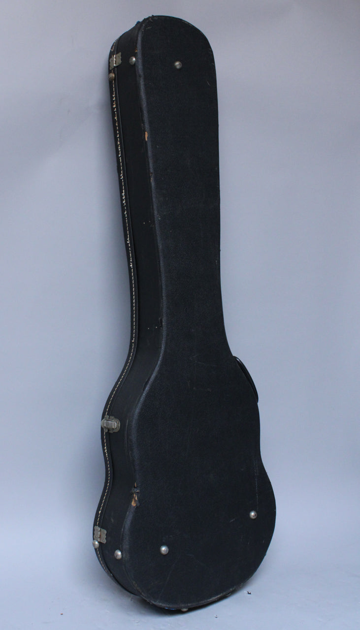 The black guitar case stands upright, showing its textured exterior, white stitching along the edge, and worn silver latches.