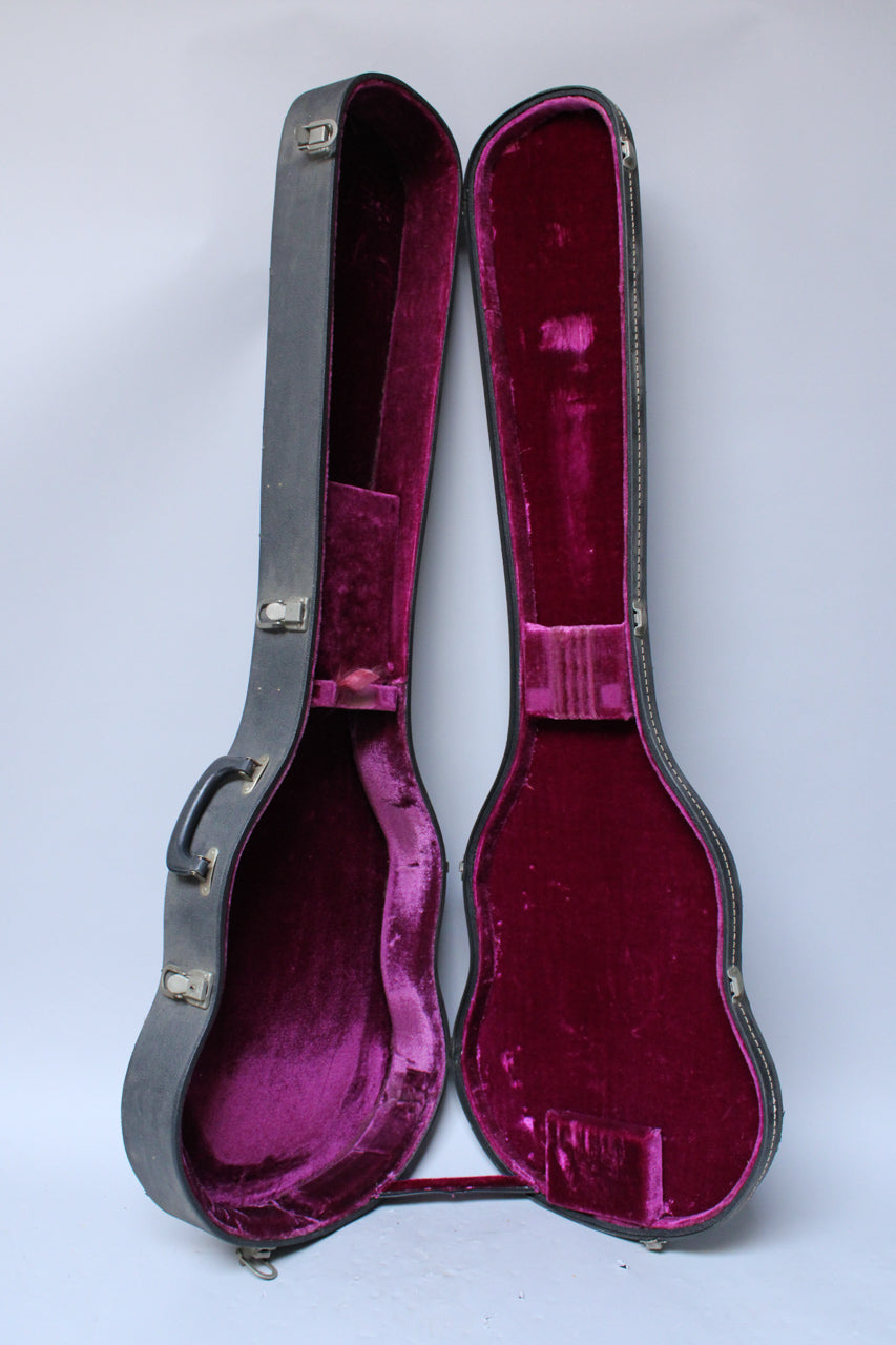 A black guitar case, open and standing upright, reveals a plush, faded purple interior with some wear.