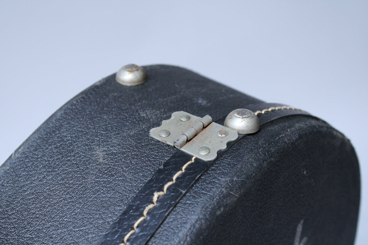 Close-up of the black textured case's top edge, showing a tarnished metal hinge and two silver dome feet, with light wear.