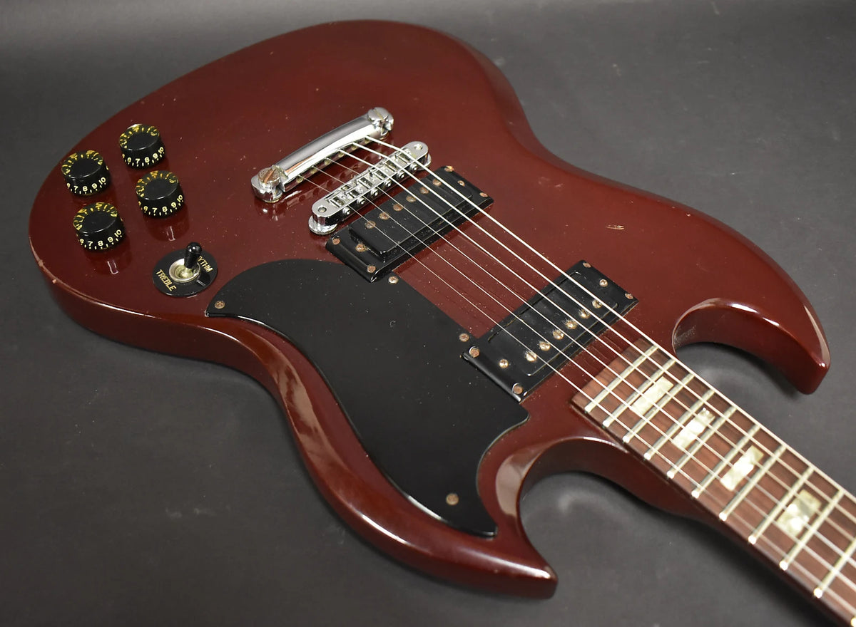 Angled view of a cherry red SG guitar body, showing the black pickguard, two humbuckers, chrome bridge, and four black control knobs.