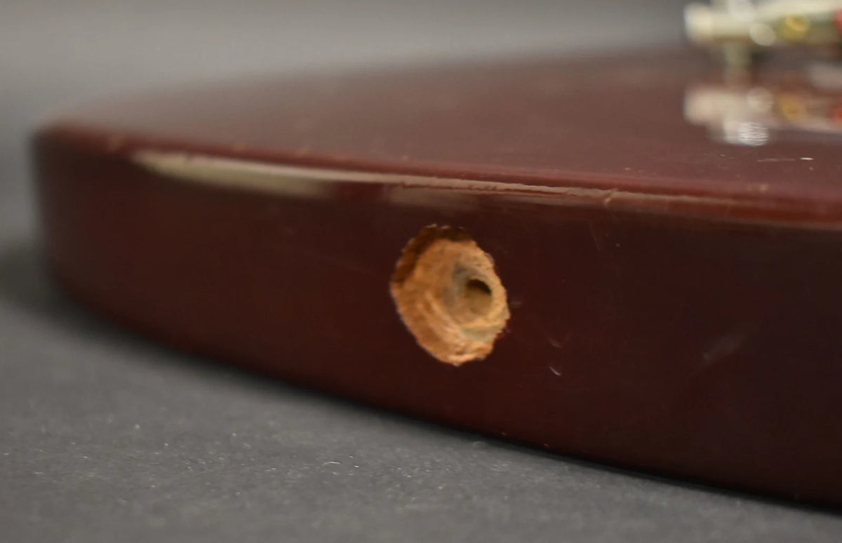 Close-up of the cherry red body's lower bout, showing a large, rough hole where the output jack should be, and finish wear along the edge.