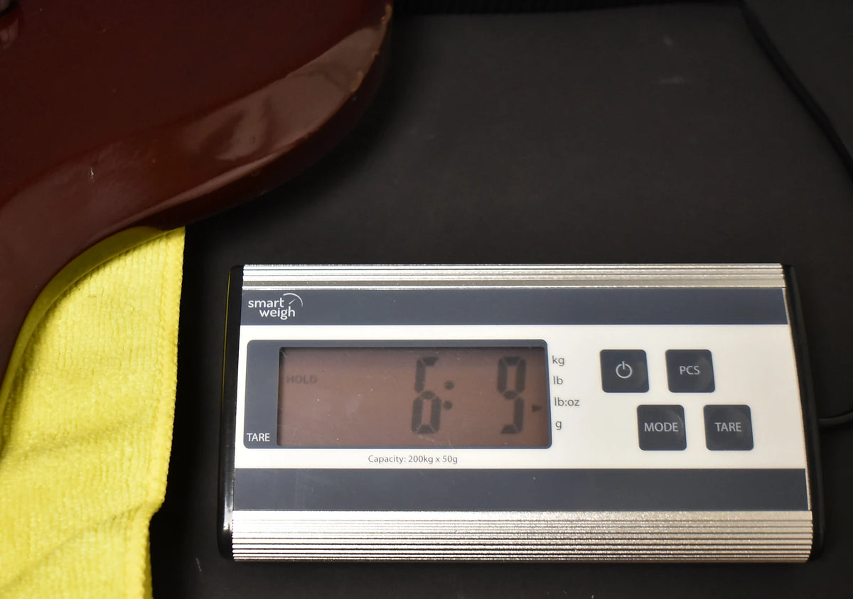 The cherry red guitar body rests on a yellow cloth, with a digital scale displaying "6:9" in pounds and ounces.