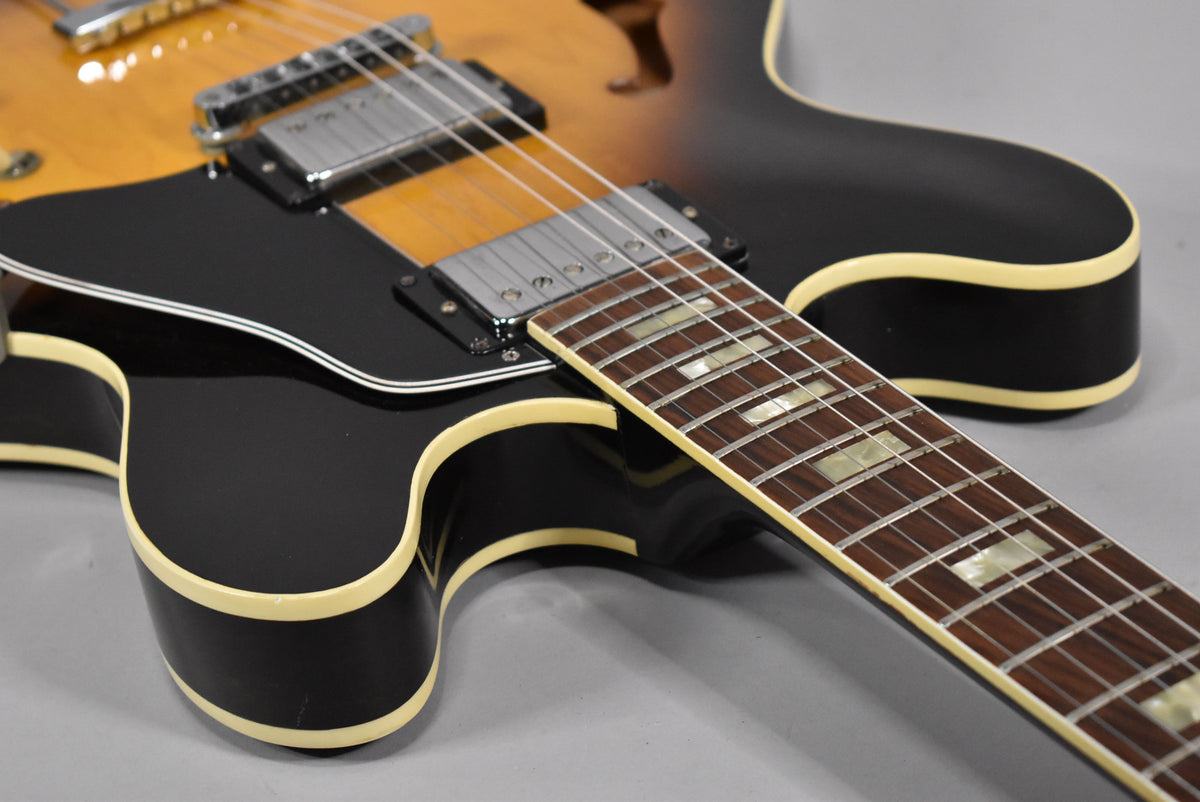 The sunburst guitar's lower bout and neck are angled from the right, showing cream binding, black pickguard, and chrome humbuckers.