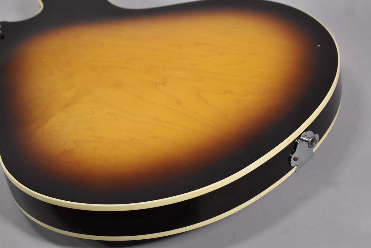 The guitar's sunburst back, showing wood grain, has a cream binding and a chrome output jack on the lower bout.