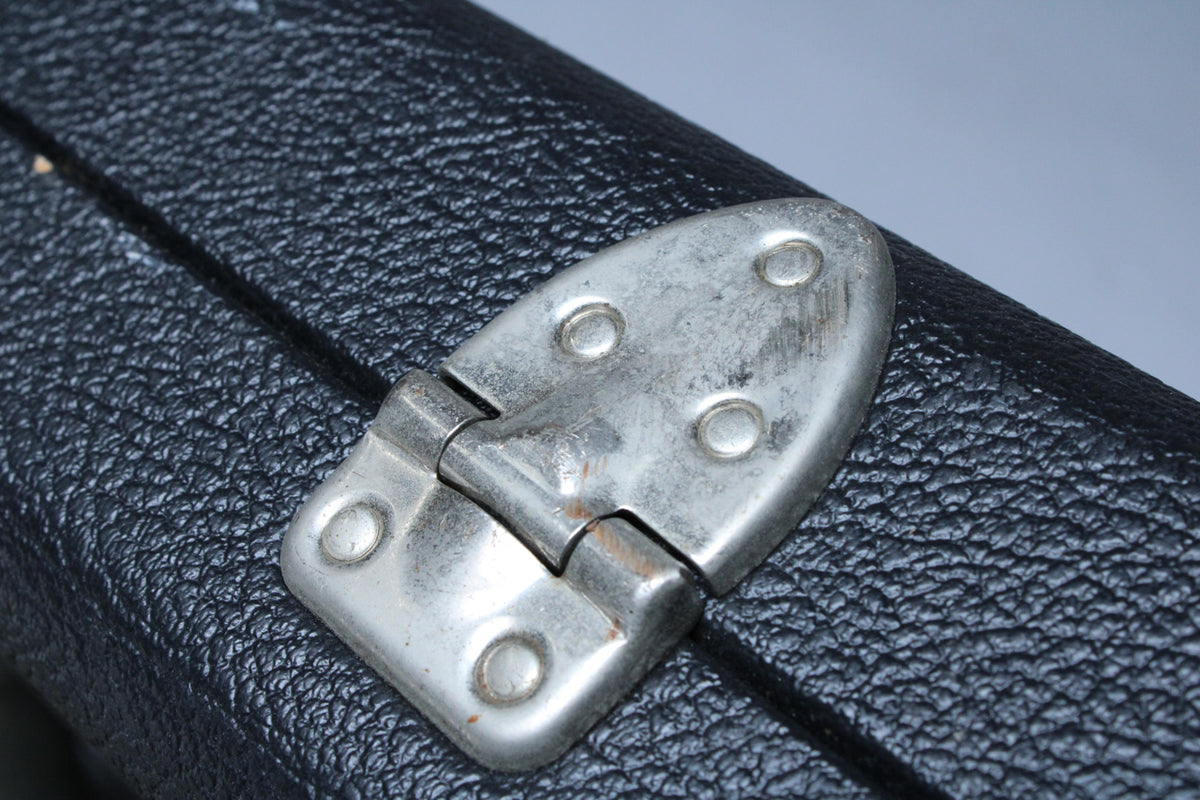 Close-up of a tarnished silver-colored hinge with visible rust on the black textured surface of the case, showing wear.