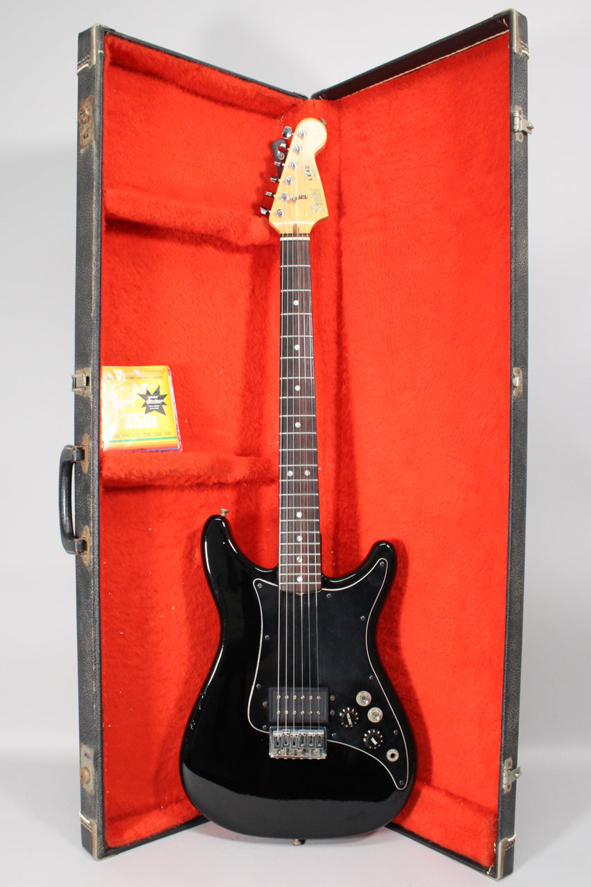A black electric guitar with a light headstock and dark fretboard, resting in an open hard case with red plush lining.