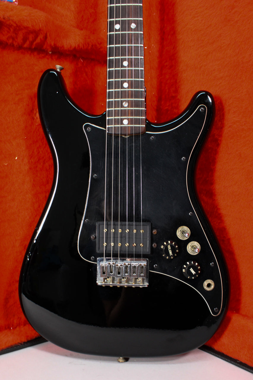 Full front view of a glossy black electric guitar with a single humbucker, three gold-capped knobs, and a white-piped black pickguard.