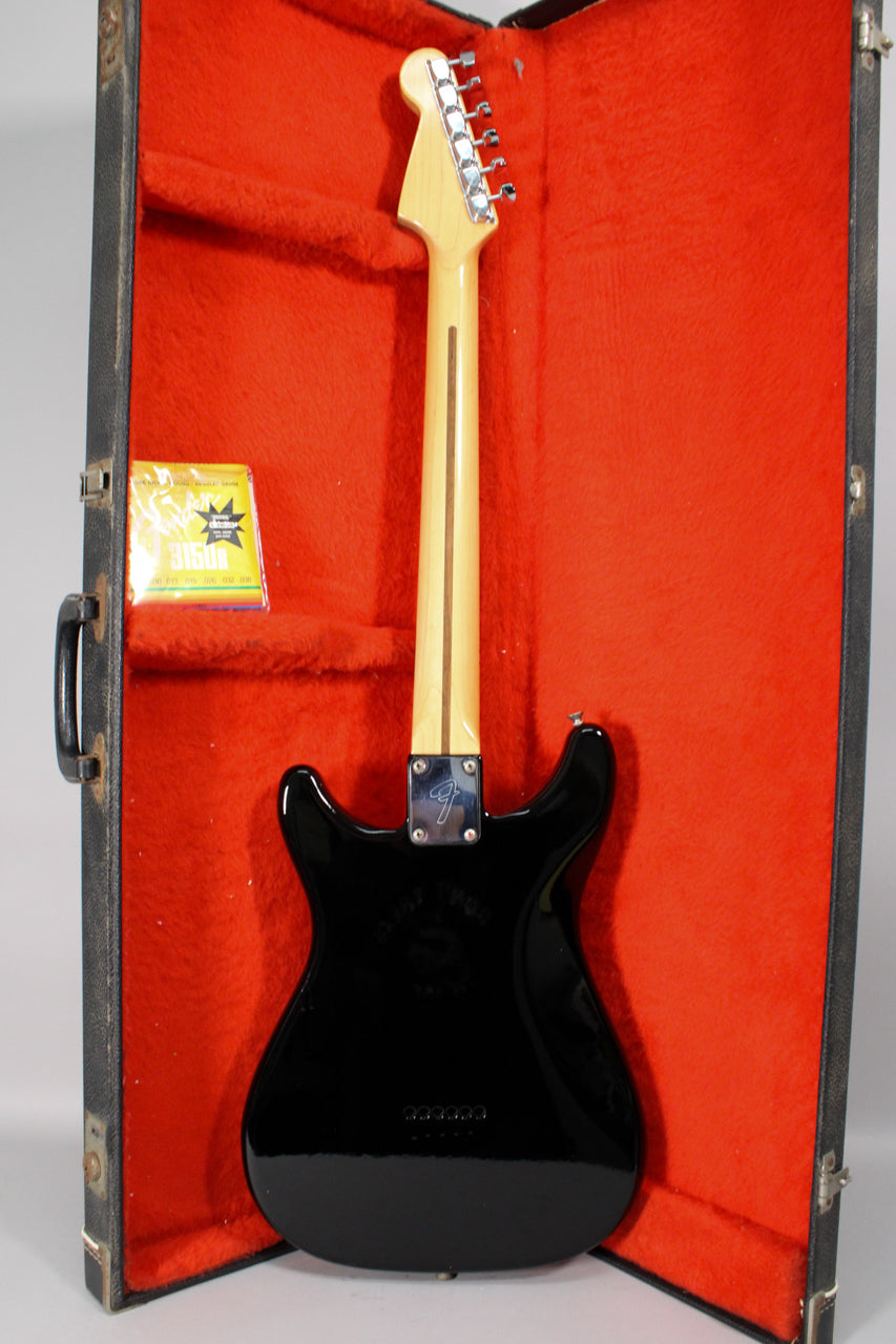 The black guitar, with a maple neck and headstock, is seen from the back in its red-lined hard case, with a pack of strings.