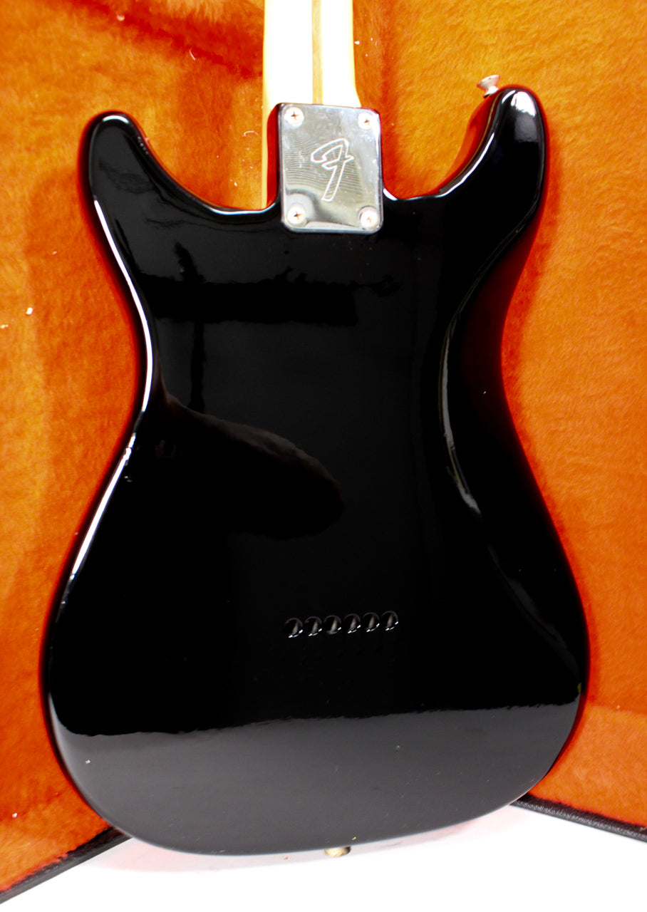 The guitar's glossy black back, with a chrome neck plate and six string ferrules, rests in an orange-lined case.