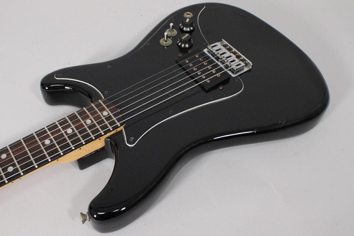 Black electric guitar, angled from the neck, showing the glossy body, single humbucker, two knobs, and three switches.
