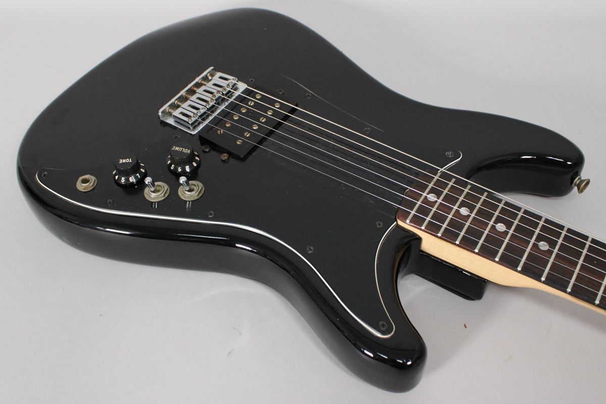 Angled view of the black guitar body, showing the chrome bridge, black humbucker, two black knobs, and three toggle switches.