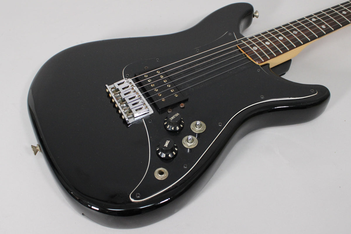 Black electric guitar, angled from above, showing the body, bridge, humbucker, two volume/tone knobs, and part of the neck.