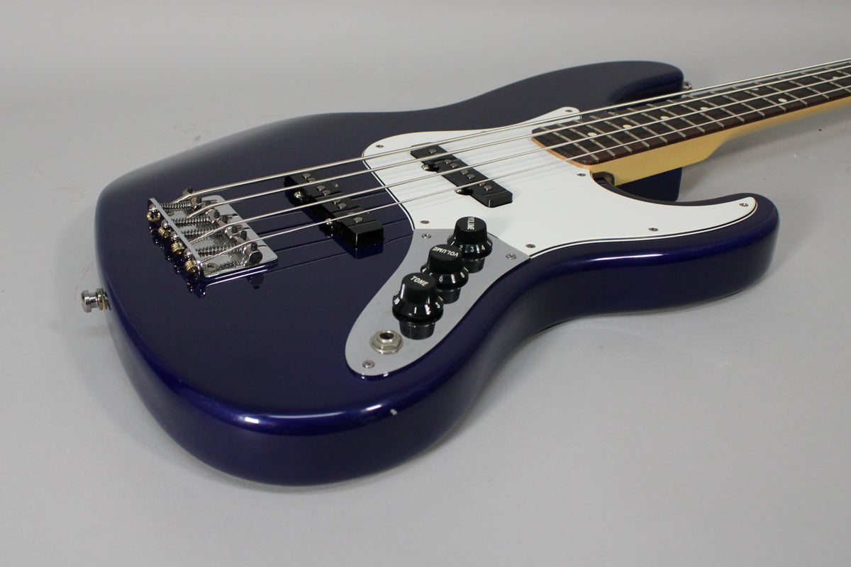 The midnight blue bass guitar is angled from the lower bout, showing the chrome bridge, white pickguard, and control plate with four black knobs.