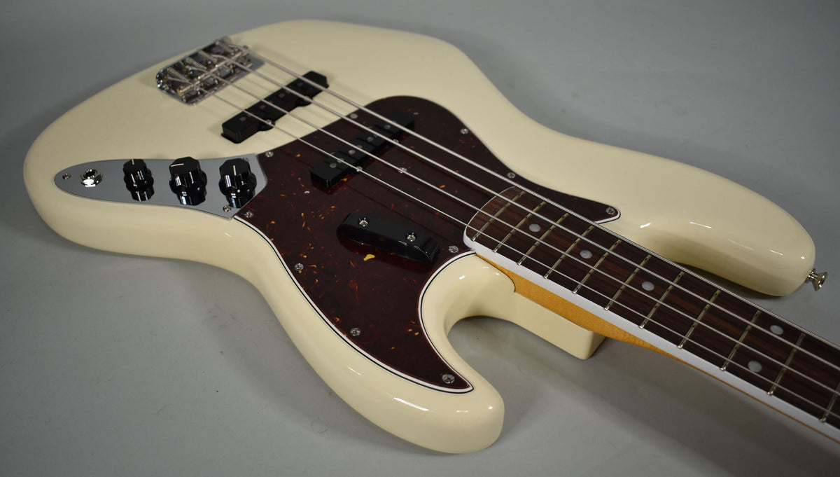 The Olympic White bass is angled from the upper left, showing its tortoise pickguard, chrome control plate with four black knobs, and rosewood fretboard.