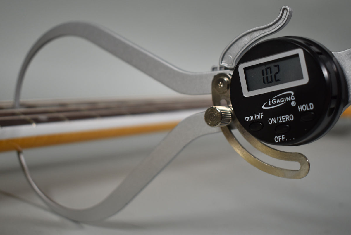 A digital caliper measures the bass neck's thickness at 1.02, showing the Olympic White finish and dark fretboard.