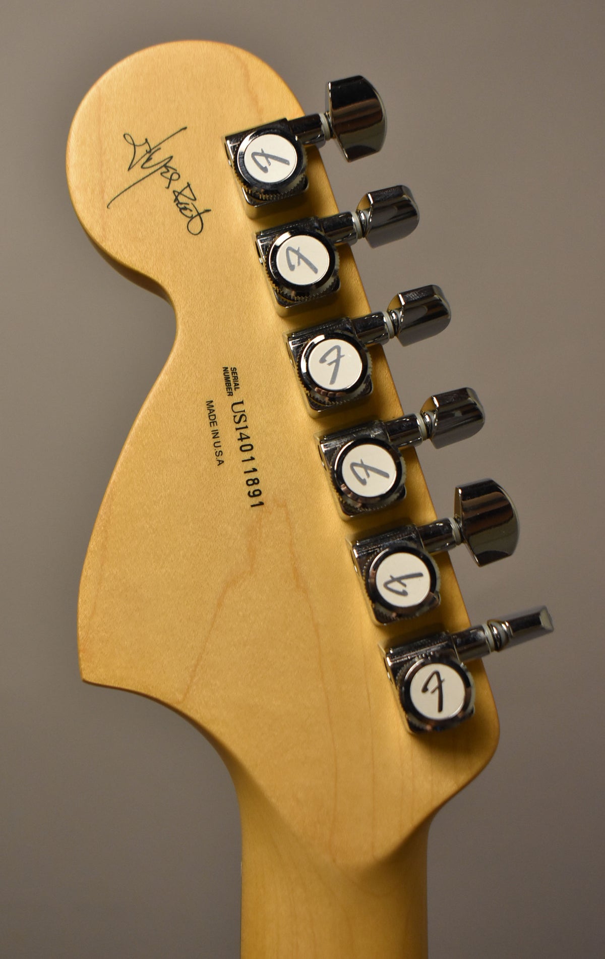 The back of a light wood headstock with six chrome tuners, a signature, and serial number.